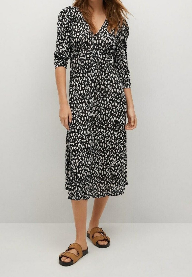 Femme portant une robe midi noire à imprimé floral blanc et manches trois-quarts, associée à des sandales à double bride couleur tan.