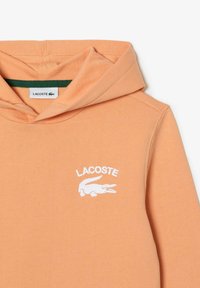 Perzikkleurige Lacoste-hoodie met een voorhoofd en een witte geborduurde krokodillenlogo en merknaam op de borst.