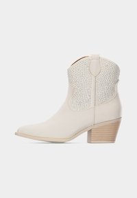 Μποτάκι αστραγάλου σε κρεμ απόχρωση με στοιβαγμένο ξύλινο τακούνι και υφασμένο εξωτερικό στρώμα με τρουκς, πλευρική θηλιά για τράβηγμα και μυτερή μύτη.