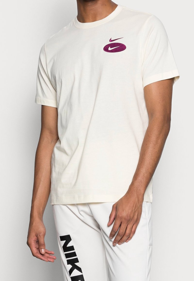 Kremowa bawełniana koszulka z krótkim rękawem, z fioletowym logo Nike na piersi. Tkanina wydaje się miękka i lekko teksturowana.