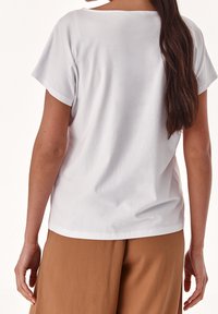 Camiseta blanca de manga corta con un escote amplio, fabricada en algodón suave, con una textura lisa y un corte relajado, combinada con pantalones beige.