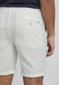 Pantalones cortos de lino claros para hombre con dobladillo enrollado, con un bolsillo trasero abotonado y una textura sutil. Diseño casual con un ajuste relajado.