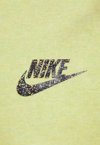 Světle žlutá tkanina s černým logem Nike, se strukturovaným barevným flekovým vzorem, umístěným ve středu a výrazným na materiálu.