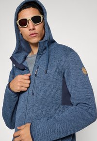 Μπλε φούτερ με φερμουάρ και υφή υφάσματος, γκρι εσωτερική επένδυση, κουκούλα με ρυθμιζόμενο κορδόνι και πλαϊνές τσέπες. Διακοσμημένο με μπαλώμα λογότυπου.