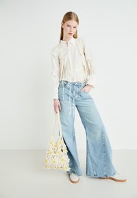 Blusa blanca texturizada con un diseño con volantes, combinada con jeans de pierna ancha de color azul claro. Sosteniendo un bolso beige con apliques florales amarillos.