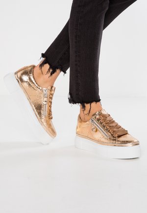 Baskets plateformes dorées métalliques avec une finition en cuir texturé, comprenant un détail zippé et des lacets en satin, associées à un jean noir effiloché.