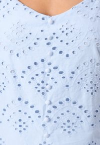 Top azul claro com padrões bordados intrincados, apresentando recortes circulares e botões decorativos ao longo da frente.