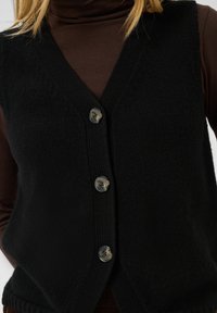 Gilet en maille noire boutonné porté sur un pull col roulé marron à manches longues, montrant le haut du torse et la zone du cou.