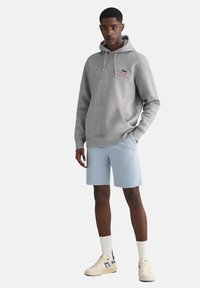 Un sweat à capuche gris avec une poche ventrale, un short bleu clair, des chaussettes blanches et des baskets crème avec des accents bleus. Le sweat porte un petit logo.