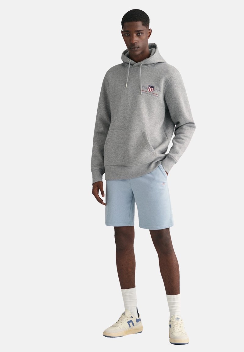 Un sweat à capuche gris avec une poche ventrale, un short bleu clair, des chaussettes blanches et des baskets crème avec des accents bleus. Le sweat porte un petit logo.
