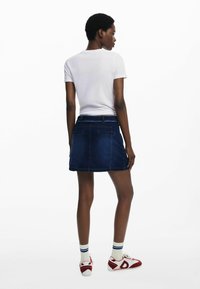 Jupe en denim bleu foncé, avec une silhouette ajustée, des poches arrière et des détails de couture. Associée à un haut à manches courtes blanc et des baskets.