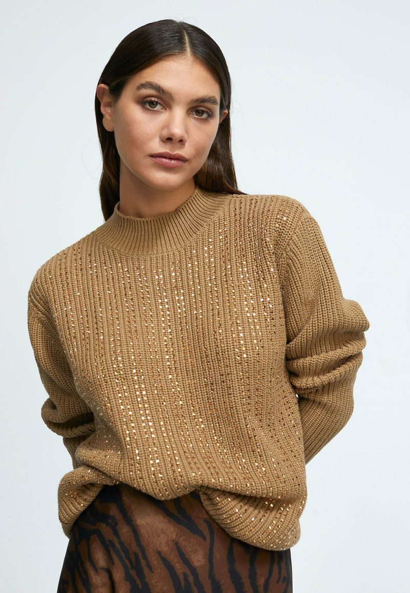 Sweater marron tricoté avec une texture côtelée et des sequins subtils, doté d'un col montant et de manches légèrement oversized, porté par-dessus un tissu à motifs.
