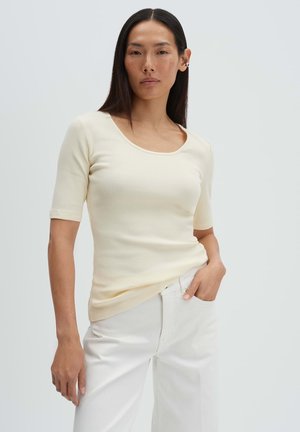 Frau mit langen schwarzen Haaren, die ein cremefarbenes, geripptes Kurzarmoberteil und hoch taillierte weiße Hosen trägt, mit einer Hand in der Tasche stehend.