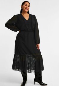 Schwarzes Langarmkleid mit V-Ausschnitt, Knopfleiste, strukturiertem Stoff und gestuftem Saum. Getragen mit knielangen schwarzen Stiefeln.