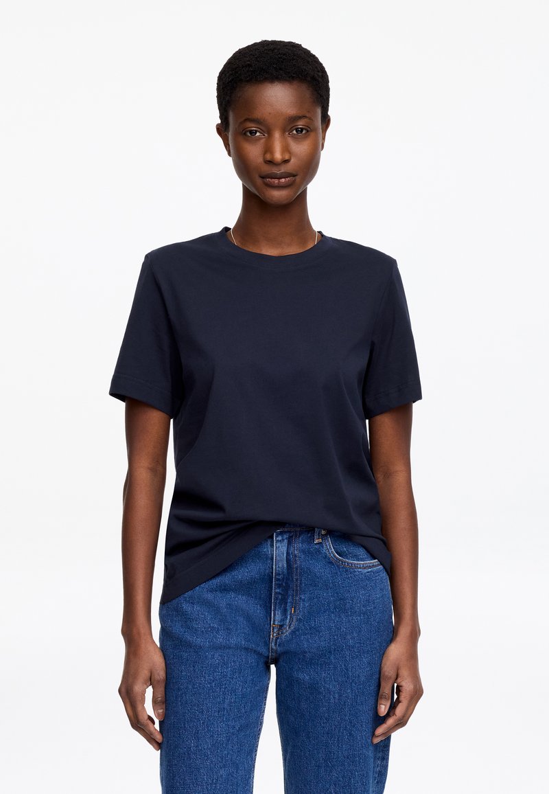 Femme aux cheveux courts portant un t-shirt uni bleu marine foncé à manches courtes, rentré dans un jean bleu, debout devant un fond blanc.