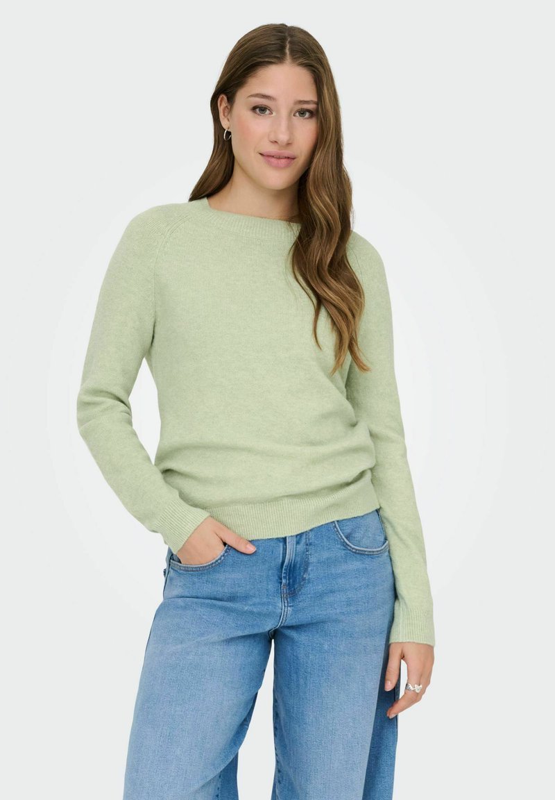 Jeune femme aux cheveux longs et ondulés, portant un pull vert clair et un jean bleu, debout avec une main dans la poche sur un fond blanc.