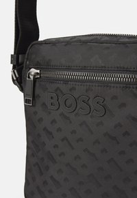Sac en bandoulière noir en textile avec un motif texturé, fermeture éclair et logo "BOSS" embossé. Sangle durable pour un transport facile.