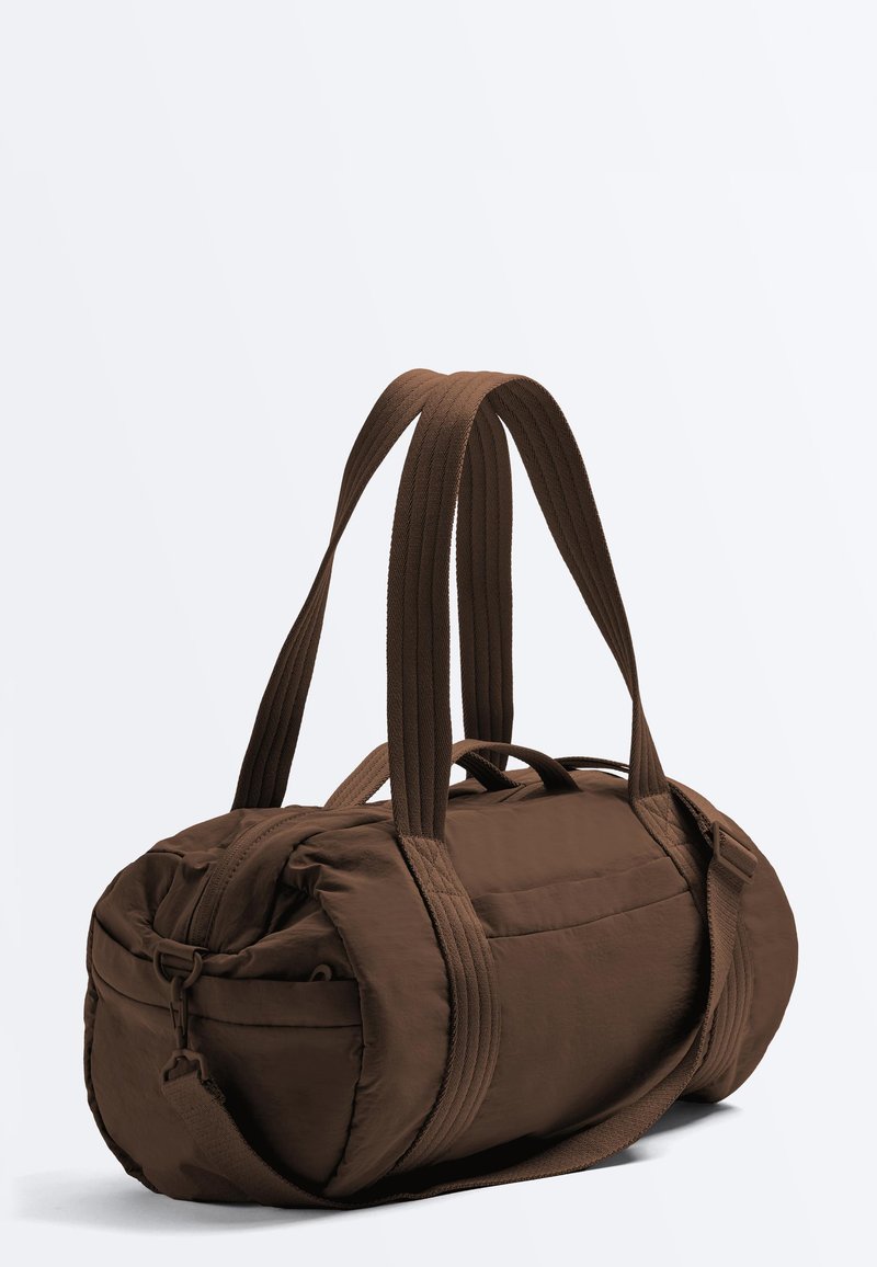 Brown cylindrical duffel bag with two long carry handles, adjustable shoulder strap, and side zippered pocket on a white background.