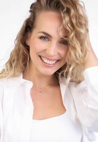 Silberne Halskette mit Wellenmuster, getragen über einem weißen Oberteil, ergänzt durch ein weißes Hemd. Das Model hat wellige blonde Haare und lächelt.