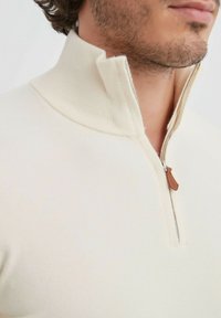 Gilet zippé couleur crème avec un col haut, présentant une fermeture éclair argentée et une languette en cuir marron. Texture douce avec une coupe ajustée.