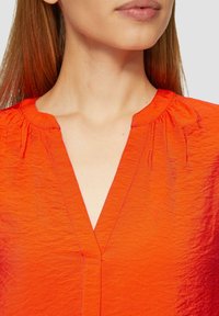 Blouse rouge, texturée, avec un décolleté en V et un detailing froncé aux épaules. Des coutures simples rehaussent l'apparence froissée du tissu.