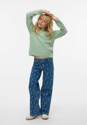 Zelena pletena majica s okruglim izrezom, u kombinaciji s plavim trapericama s cvjetnim uzorkom. Model nosi svijetle tenisice.