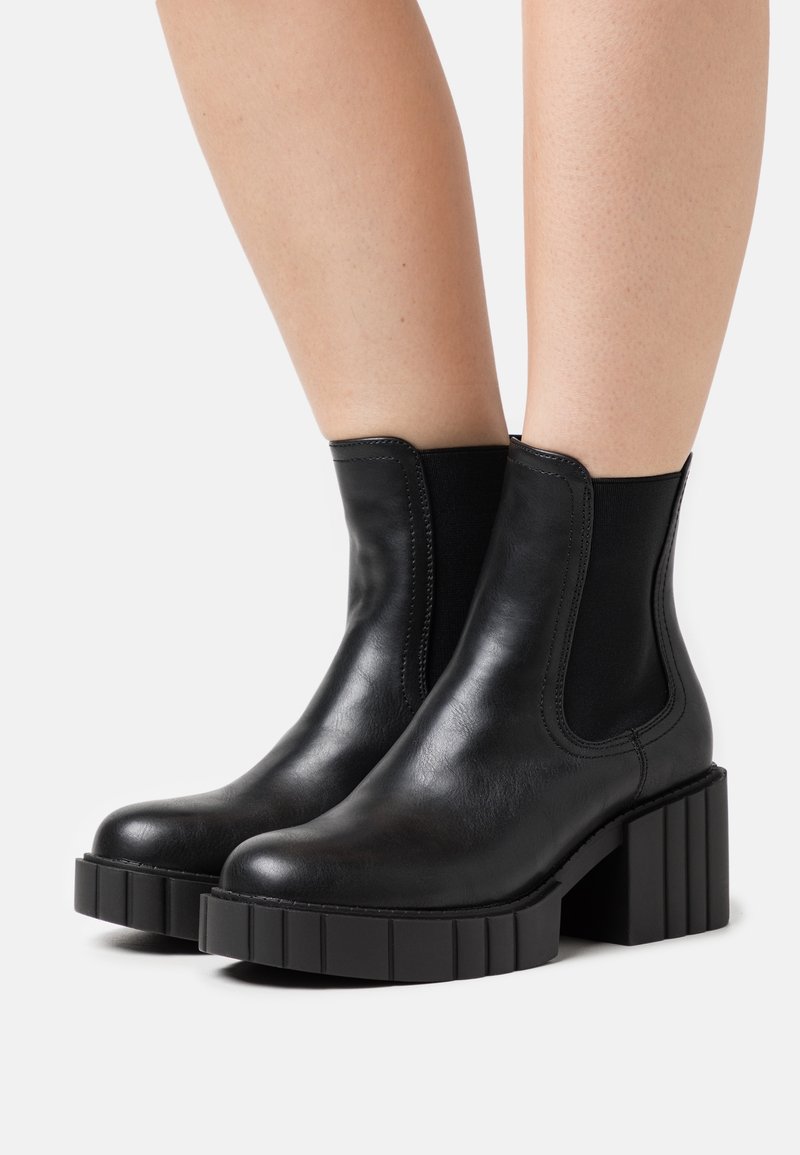 Bottes basses en cuir noir avec talons carrés épais et semelles à plateforme rainurées portées sur des jambes à peau claire sur un fond blanc.