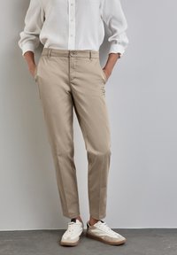 Chinos beige con una vestibilità tailored, caratterizzati da tasche frontali e chiusura con bottone, abbinati a sneaker bianche e una camicia bianca ampia.