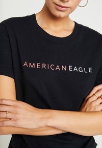 Svart t-shirt med korta ärmar, med texten "AMERICANEAGLE" i gradientfärger; avslappnad passform med ribbat halsringning och mjukt tyg.