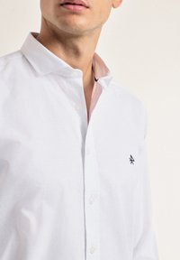 Camisa blanca de botones con un cuello suave, forro interior sutil en rosa y un pequeño logotipo en navy en el lado izquierdo del pecho. Tejido suave y transpirable.