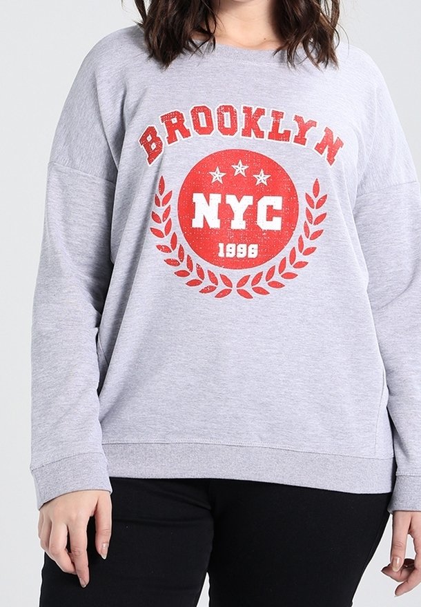 Sweat-shirt gris avec l'inscription « BROOKLYN NYC 1996 » en rouge et blanc entourée d'une couronne de laurier et d'étoiles, porté par une personne aux cheveux foncés.