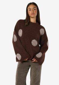 Ung kvinna som bär en brun fluffig tröja med stora beiga prickar och grå kordelbyxor, stående med händerna vid midjan.