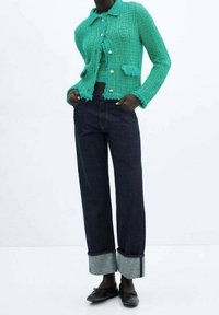 Türkis geflochtene Jacke mit fransigen Kanten und goldenen Knöpfen, kombiniert mit dunkelblauen weit geschnittenen Jeans mit umgeschlagenen Säumen und blauen Nähten.