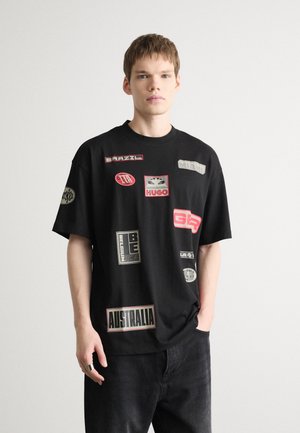 T-shirt noir oversize avec divers patchs, y compris du texte et des logos en rouge, blanc et gris. Col rond, manches courtes et coupe décontractée.