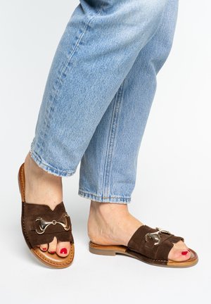 Nohy v hnědých semišových sandálech na nazouvání se stříbrnými kovovými doplňky a červeným lakem na nehtech, ve spojení se světle modrými džínami.