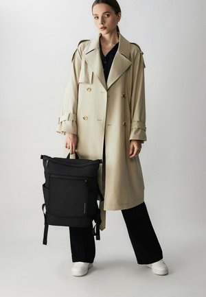 Cappotto beige con bottoni dorati doppiopetto, camicia nera, pantaloni neri a gamba larga e uno zaino nero con cerniere e cinturini.