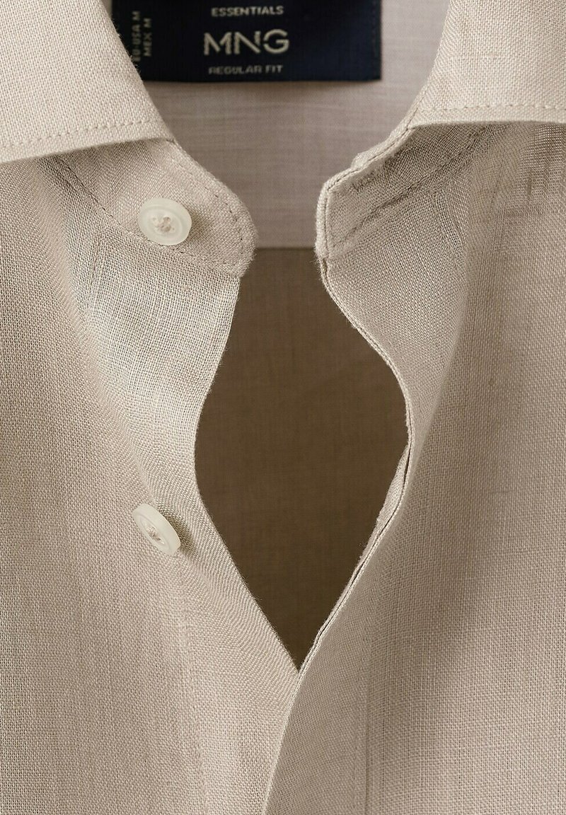 Chemise en lin beige clair avec col ouvert, deux boutons blancs défaits, et une étiquette sombre indiquant "ESSENTIALS MNG REGULAR FIT".
