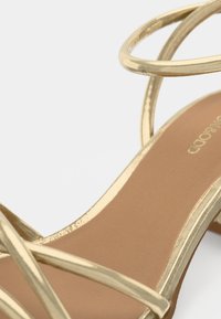 Sandale aurii cu barete metalice curbate, talpă interioară netedă de culoare tabac și toc scăzut, evidențiind un design minimalist și un finisaj elegant.