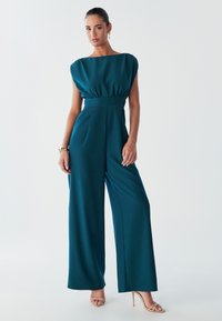 Model wearing a teal jumpsuit with a fitted waist and wide legs, styled with a bracelet and strappy high heels, against a light background.