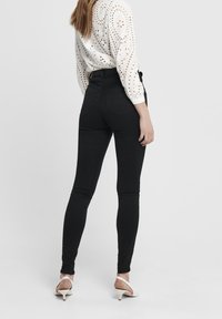 Jeans ajustados negros con bolsillos traseros, de cintura alta y tela elástica, combinados con una blusa blanca de encaje y sandalias de tacón blancas.