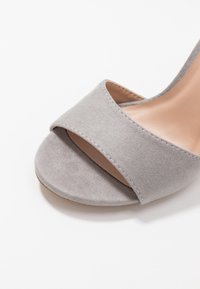 Sandale ouverte en daim gris clair avec large bride avant et semelle intérieure lisse de couleur tan sur fond blanc.