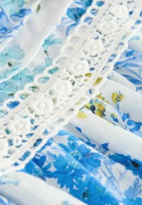 Blommönstrad tyg med blå och vita mönster och inslag av gult. Dekorativ vit spetskant längs den vertikala sömmen. Mjuk, lätt textur.