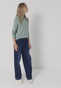 Hellgrünes Langarmoberteil, kombiniert mit locker sitzenden dunkelblauen Jeans und weißen Sneakern, steht vor einem neutralen Hintergrund.