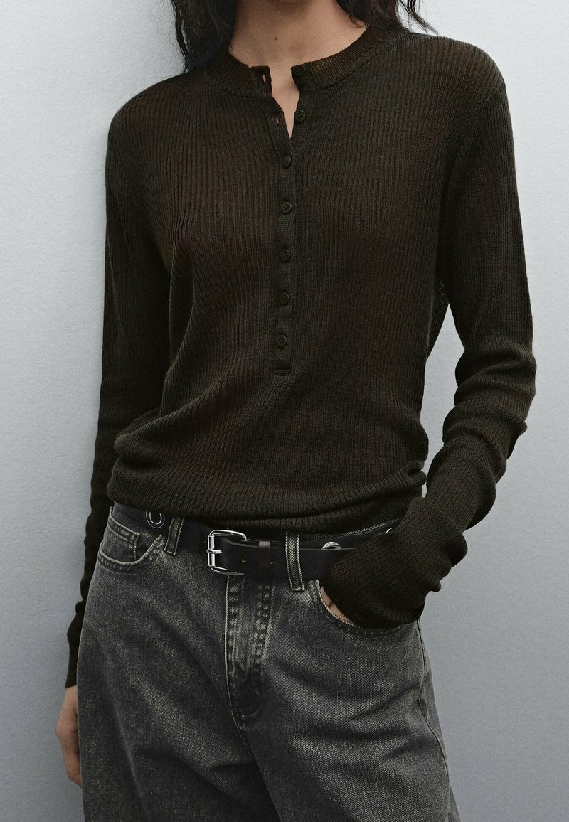 Femme portant un haut henley à manches longues côtelé de couleur foncée, rentré dans un jean noir délavé taille haute avec une ceinture noire et une main dans la poche.