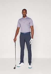 Golfový outfit zahrnující světle fialovou polo košili s límečkem, námořnické kalhoty, modrý opasek, bílé boty a držící golfovou hůl.