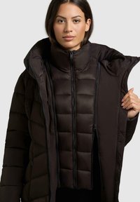 Giacca a vento nera con colletto alto e numerosi zip, con una texture trapuntata. Lo strato interno è anche un gilet imbottito nero.