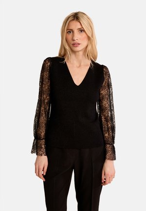 Donna con capelli biondi che indossa un top nero con scollo a V e maniche lunghe in pizzo trasparente e pantaloni neri, in piedi davanti a uno sfondo chiaro e uniforme.