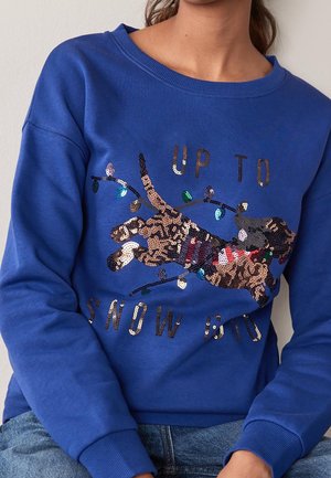 Sweat-shirt bleu avec un teckel en sequins portant une écharpe et des guirlandes lumineuses, avec le texte « UP TO SNOW GOOD » en sequins.