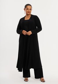 Zwarte lange cardigan met manchetten versierd met kralen, gemaakt van zachte stof. Draagt over bijpassende top en wijde broekspijpen. Open voorkant ontwerp.