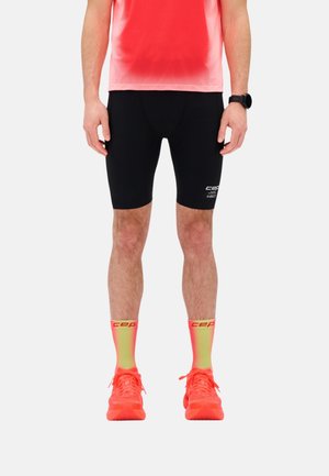 Mann trägt schwarze Kompressionsshorts, neon-gelbe und rote CEP-Socken, rote Laufschuhe und ein rot verlaufendes Sportshirt, steht vor weißem Hintergrund.